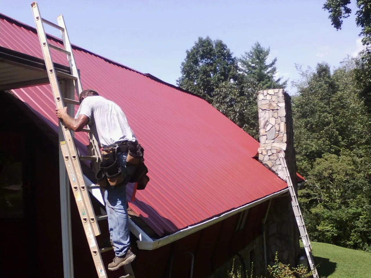 Roofer on a ladder beside a red metal roof and stone chimney — Soffit & Fascia Repair by Roofing Brothers in Humansville